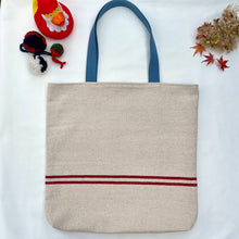 Load image into Gallery viewer, Tote bag. Heavyweight natural woven canvas with two horizontal red stripes. Lined with an ex-designer striped cotton shirting fabric.