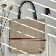 Load image into Gallery viewer, Tote bag. Heavyweight natural woven canvas with two horizontal red stripes. Lined with a beautiful striped cotton shirting fabric.
